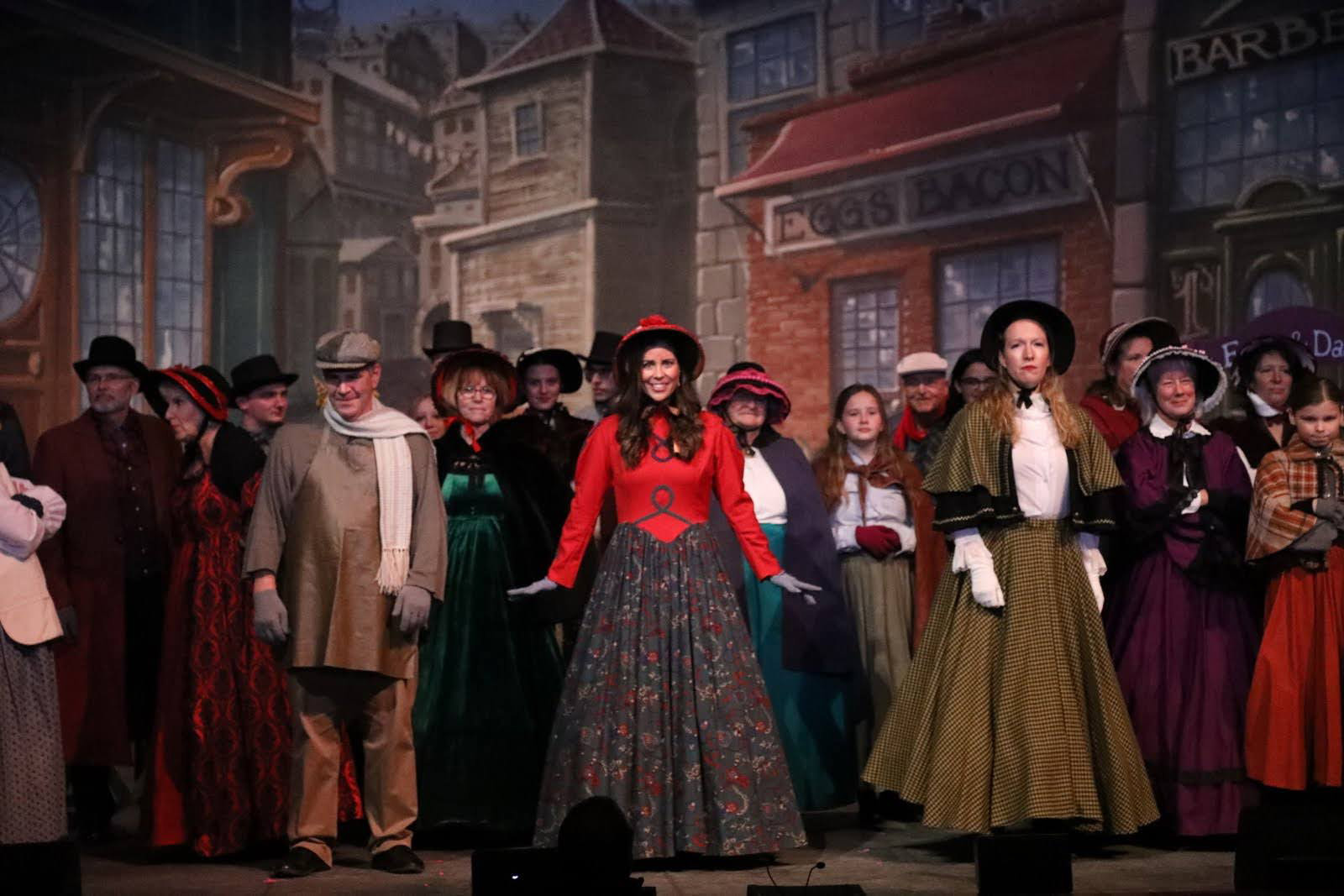 Men, women and children in Dickens costumes gather in a town square scene.
